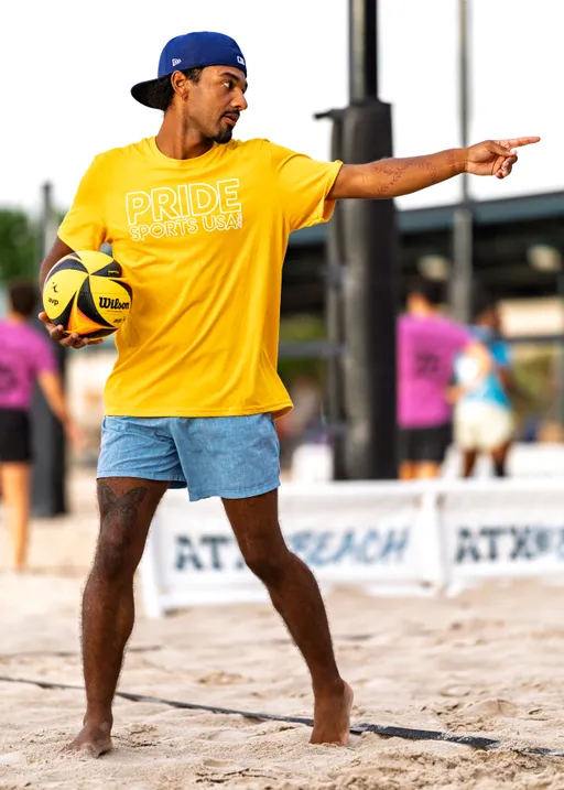 A volleyball player wearing a yellow "Pride Sports USA" t-shirt, blue shorts, and a blue cap stands barefoot on the sand while holding a volleyball and pointing toward the court, with other participants and volleyball nets visible in the background.