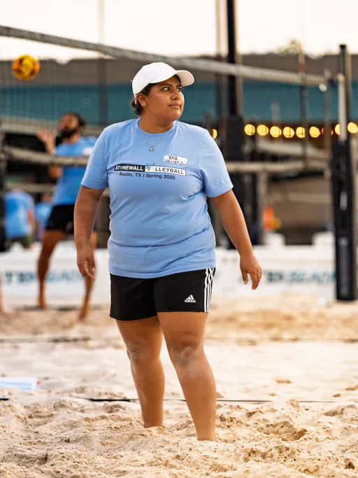 A volleyball player wearing a light blue Stonewall Volleyball Spring 2025 t-shirt, black shorts, and a white baseball cap stands on a sandy court, with another player in the background preparing to hit a volleyball over the net.