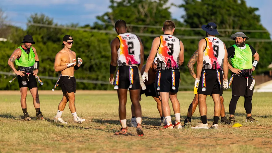 ATX Heat (Black) players huddle on a sunlit grass flag‑football field during practice for Gay Bowl 2025, with cones marking the drill. Athletes wear white jerseys with colorful splashes and numbers (e.g., 2, 3, 9), black shorts, and flag belts; some have neon green pinnies, gloves, caps, or wide‑brim hats as teammates listen and prepare for the next play.