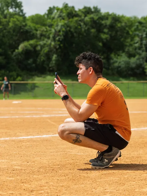 A person wearing an orange shirt, black shorts, athletic shoes, and a watch is squatting on a kickball field, holding up a smartphone toward the playing area, with trees and grass in the background.