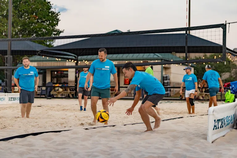 On a sand volleyball court, a player in a blue team shirt crouches low at the sideline to dig a yellow-and-black ball while teammates in matching shirts watch and ready themselves near the net; the outdoor facility features a pavilion with string lights and “ATX Beach” branding on the court barrier.