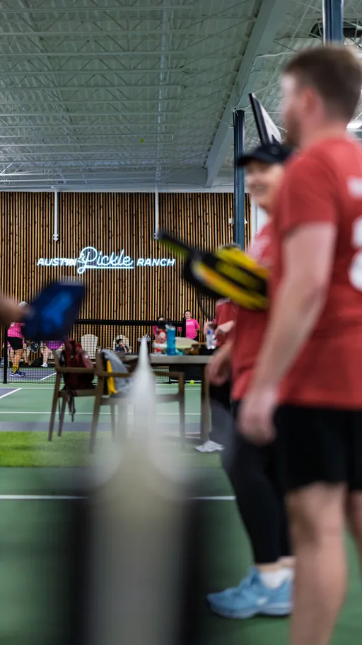 Players in red shirts stand on an indoor pickleball court, holding paddles and appearing engaged in conversation or preparing for a game, with other participants and people visible in the background near tables and a wood-paneled wall.