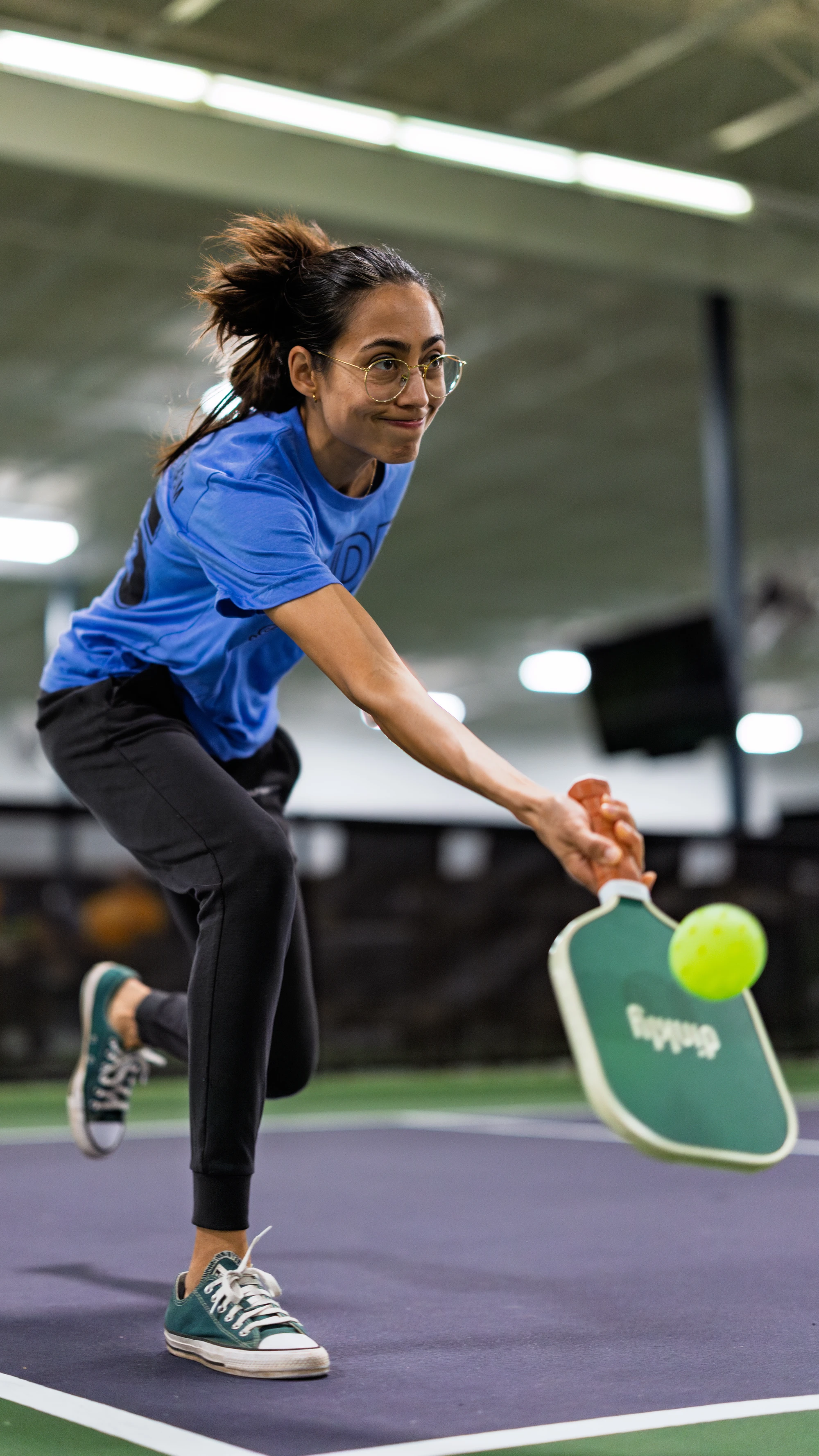 A pickleball player in athletic clothing and glasses lunges forward on an indoor court, swinging a paddle to hit a bright yellow pickleball.