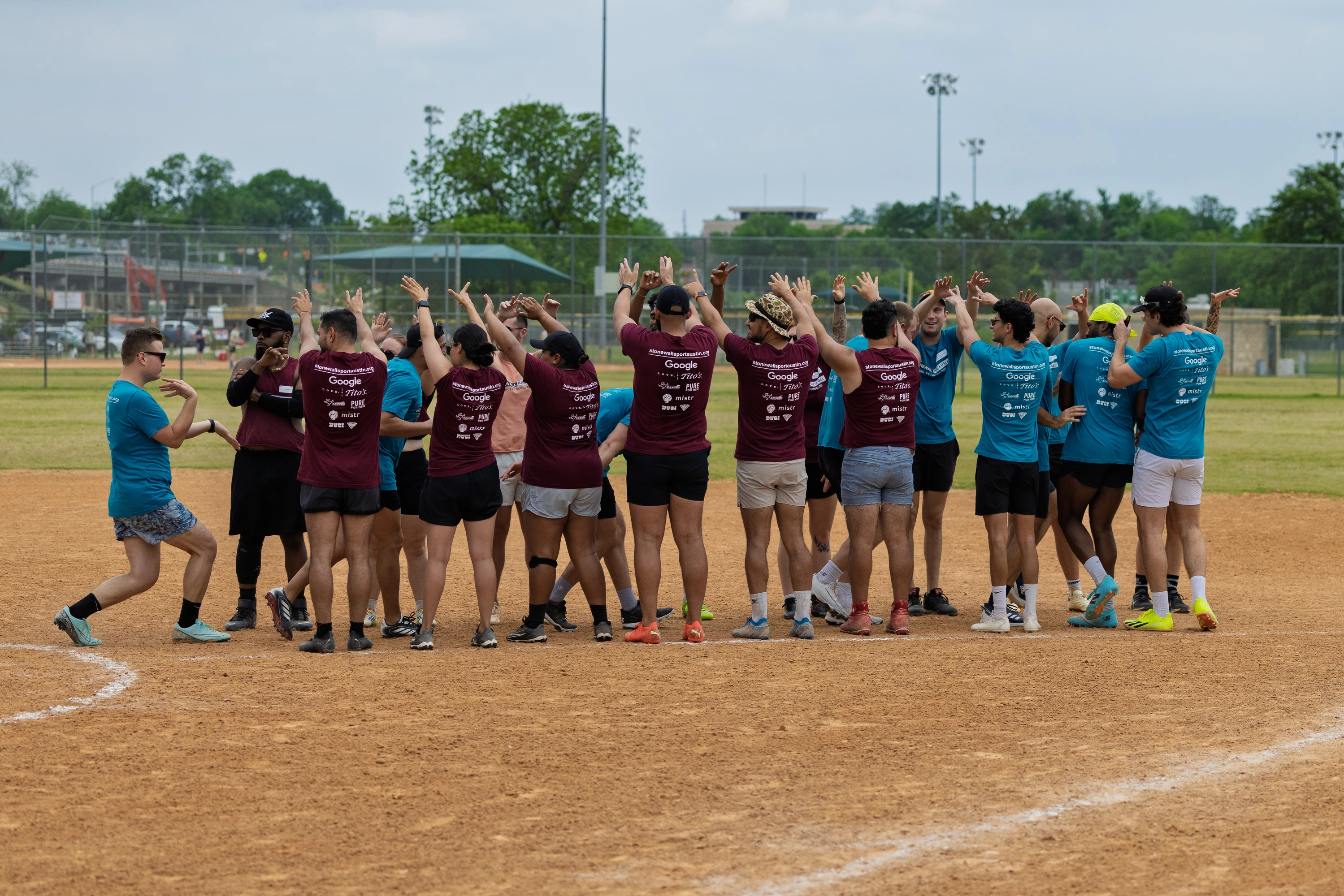 Players wearing maroon and teal team shirts are gathering in a circle on a kickball field, raising their arms and celebrating together after a game. The field is surrounded by grass, fencing, and distant sports facilities.