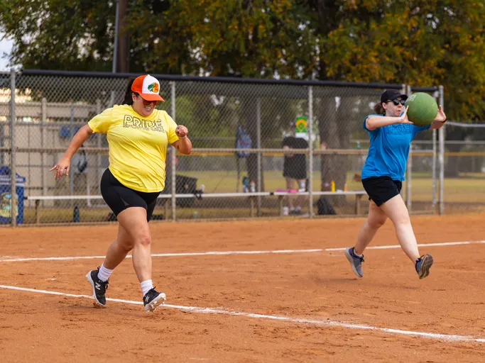 Action shot on a kickball field: a player in a yellow PRIDE shirt and orange trucker hat sprints along the baseline while a defender in a blue shirt, black cap, and sunglasses lunges forward to throw a green kickball. Dusty infield with a chain-link fence and trees in the background.