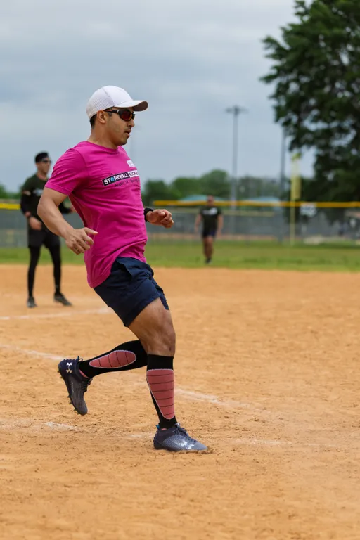 A kickball player wearing a bright pink shirt, dark shorts, tall pink socks, and sunglasses is stepping forward on a sandy field, appearing to be in motion during gameplay. Other players and a grassy outfield are visible in the background.