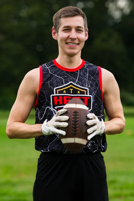 An ATX Heat (Orange) player stands on a grassy field, smiling and holding a football with white receiver gloves at chest level. The athlete wears a black sleeveless ATX HEAT jersey with red-orange trim, practicing outdoors for Gay Bowl 2025.