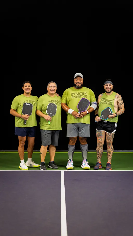 Four pickleball players stand side by side on an indoor pickleball court, each holding a paddle and wearing matching green "Pride Pickleball" shirts, with two players wearing wristbands and one wearing a cap. The group is smiling and posing for a team photo, and one player has visible tattoo sleeves and athletic shorts.