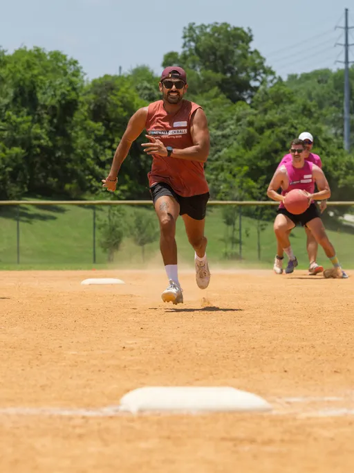 A player in a sleeveless shirt, shorts, and cap sprints on a kickball field, while another player in a pink shirt chases after a rolling kickball in the background. The scene takes place outdoors on a sunny day with trees and grassy areas visible beyond the field.
