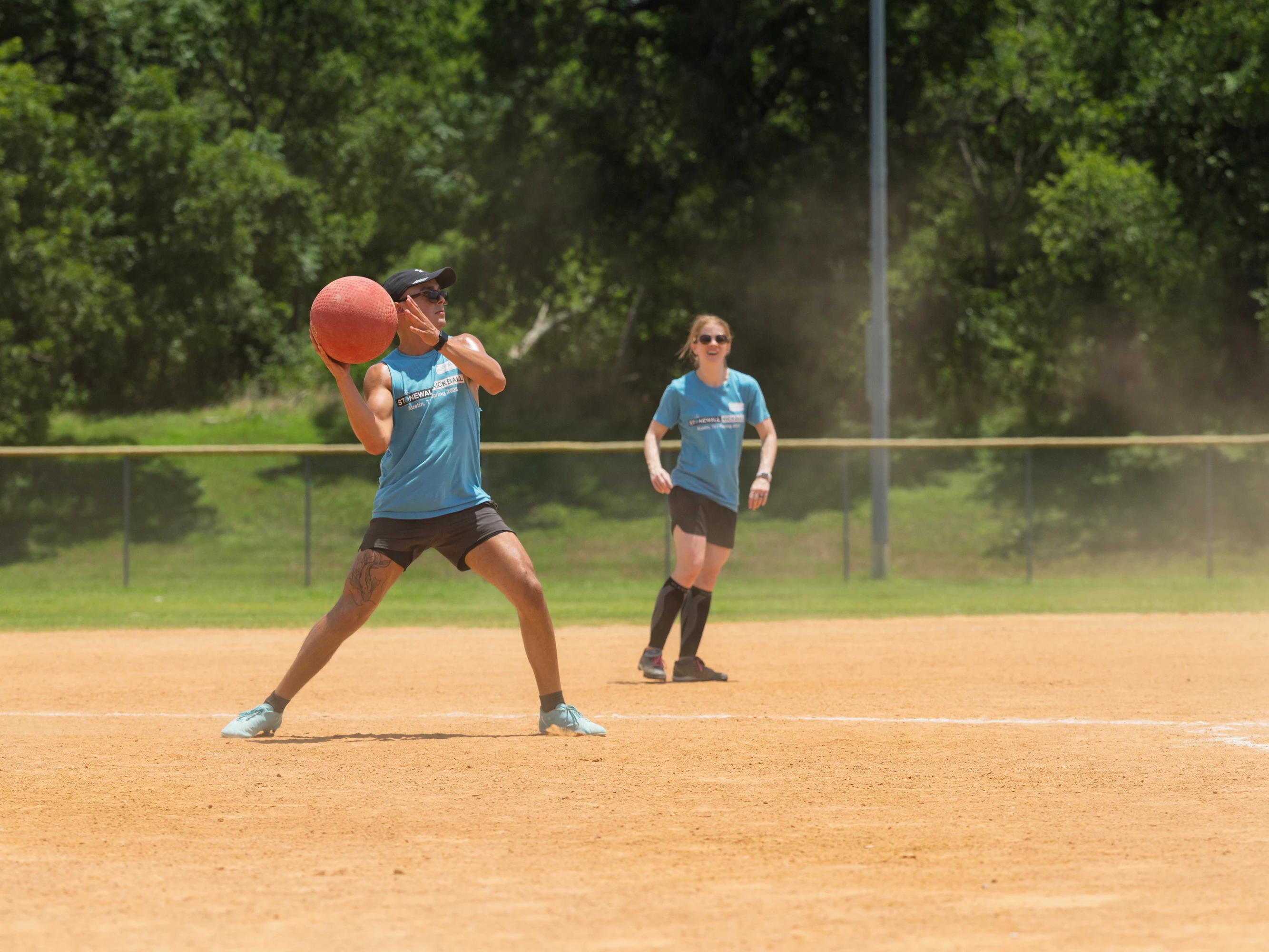 A kickball player in a blue jersey and visor is preparing to throw a red kickball on a dusty field, while another player in similar attire stands alert in the background.