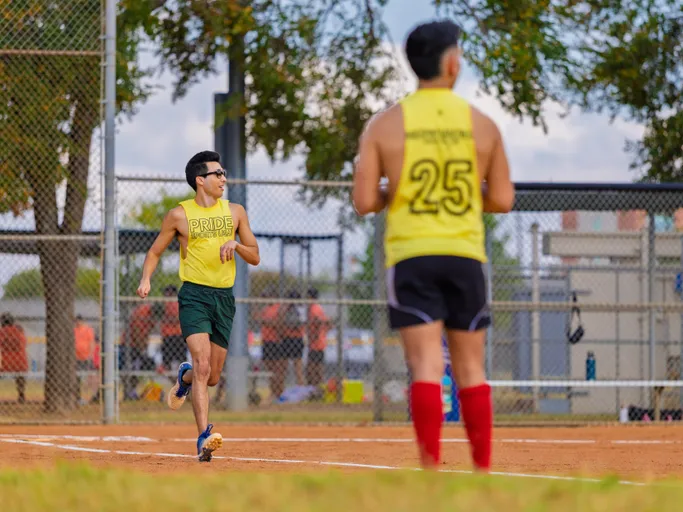 On a sunny park kickball field, two Pitch Please players in yellow jerseys compete: a runner in green shorts and sunglasses sprints up the baseline while a teammate with jersey number 25 and red knee-high socks watches near the infield, with chain-link fencing and trees in the background.