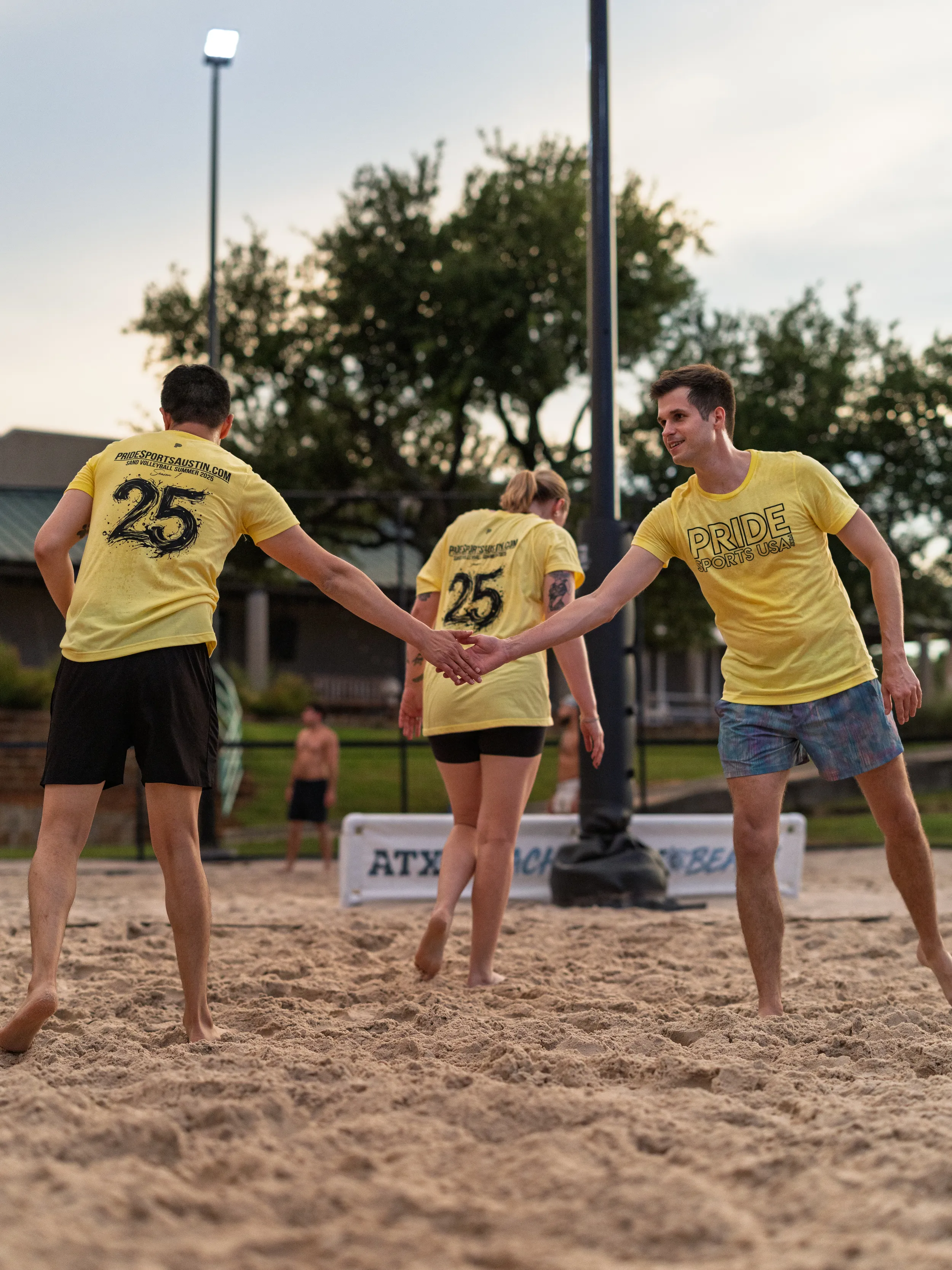 Several sand volleyball players in yellow PRIDE team shirts—“PRIDE” on the front and “25” on the back—exchange a low-five as they cross paths on the sand. The evening scene is lit by a tall lamp; players are barefoot in shorts, with trees, houses, and an “ATX” banner in the background.