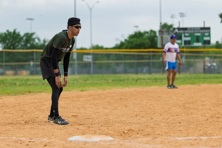 A player in a black headband, dark shirt, and leggings is positioned alertly near a base on a kickball field, while another person stands in the outfield wearing a white and purple shirt and blue shorts.