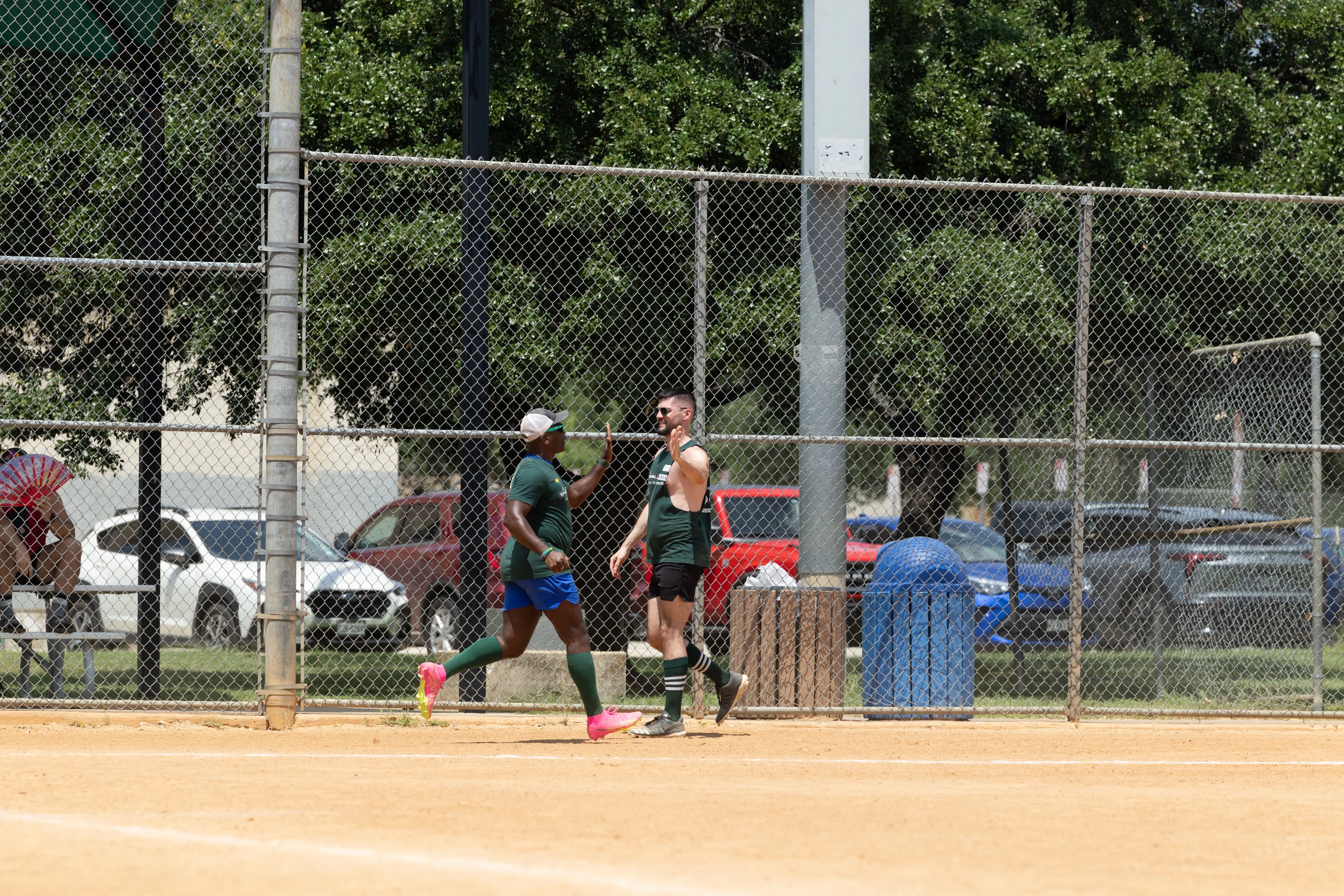 Two kickball players, both wearing dark green jerseys and knee-high socks, are giving each other a high five on the dirt field near a chain-link fence, with parked cars and trees visible in the background. One player is also wearing blue shorts, a white hat, and bright pink shoes.