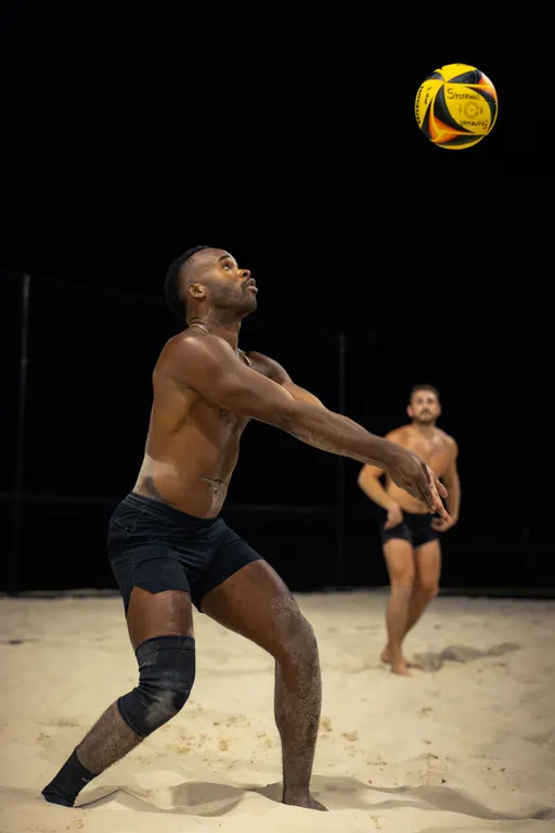 On a night-lit sand volleyball court, a player in black shorts and a knee brace prepares to bump a yellow-and-red volleyball, while a teammate in dark shorts watches and readies behind.