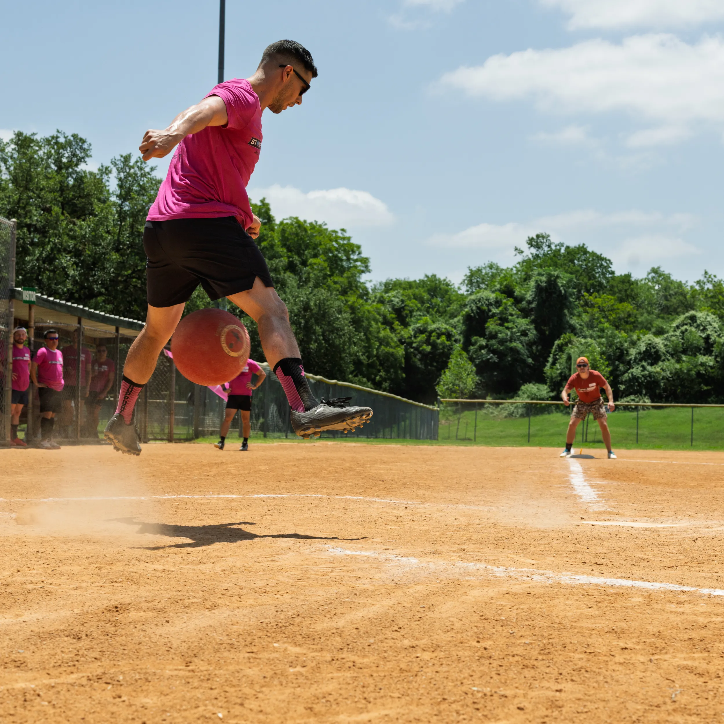 A player in a bright pink team shirt and black shorts leaps to kick a red kickball, kicking up dust on a sunny kickball field with white chalk baselines. Teammates in matching pink shirts watch from the dugout as a fielder readies near the line in the outfield.