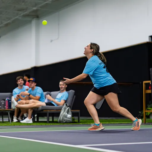 A pickleball player in mid-motion prepares to hit a ball, wearing a blue athletic shirt, black shorts, and sneakers, on an indoor court. Several people in matching blue shirts watch from benches in the background.