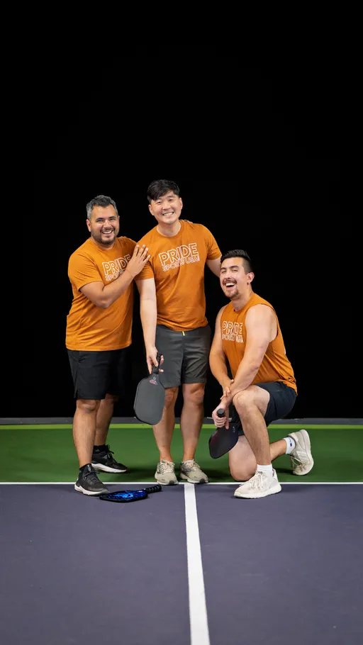 Three pickleball players wearing matching orange "Pride Pickleball" shirts pose together on an indoor court, each holding a pickleball paddle, with one player kneeling and the others standing beside them. A paddle and ball rest on the court in front of the group.