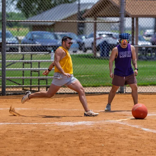 A player in a yellow tank top and gray shorts is kicking a red kickball on a dirt field, while another player wearing a purple "PRIDE KICKBALL LEAGUE" tank top, shorts, and a blue bandana observes nearby.