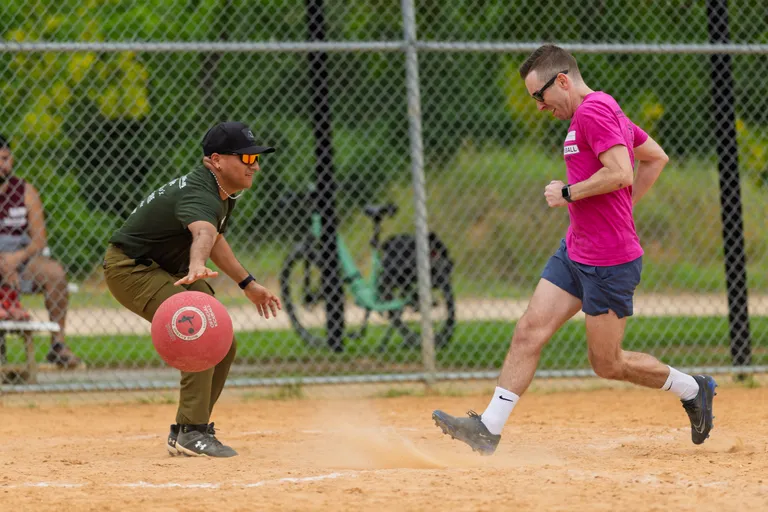 Two kickball players are in action on a dirt field; one in a green shirt and dark cap is reaching for the ball, while another in a bright pink shirt and athletic shoes is running past, seemingly attempting to score or avoid being tagged. A chain-link fence and some bicycles are visible in the background.