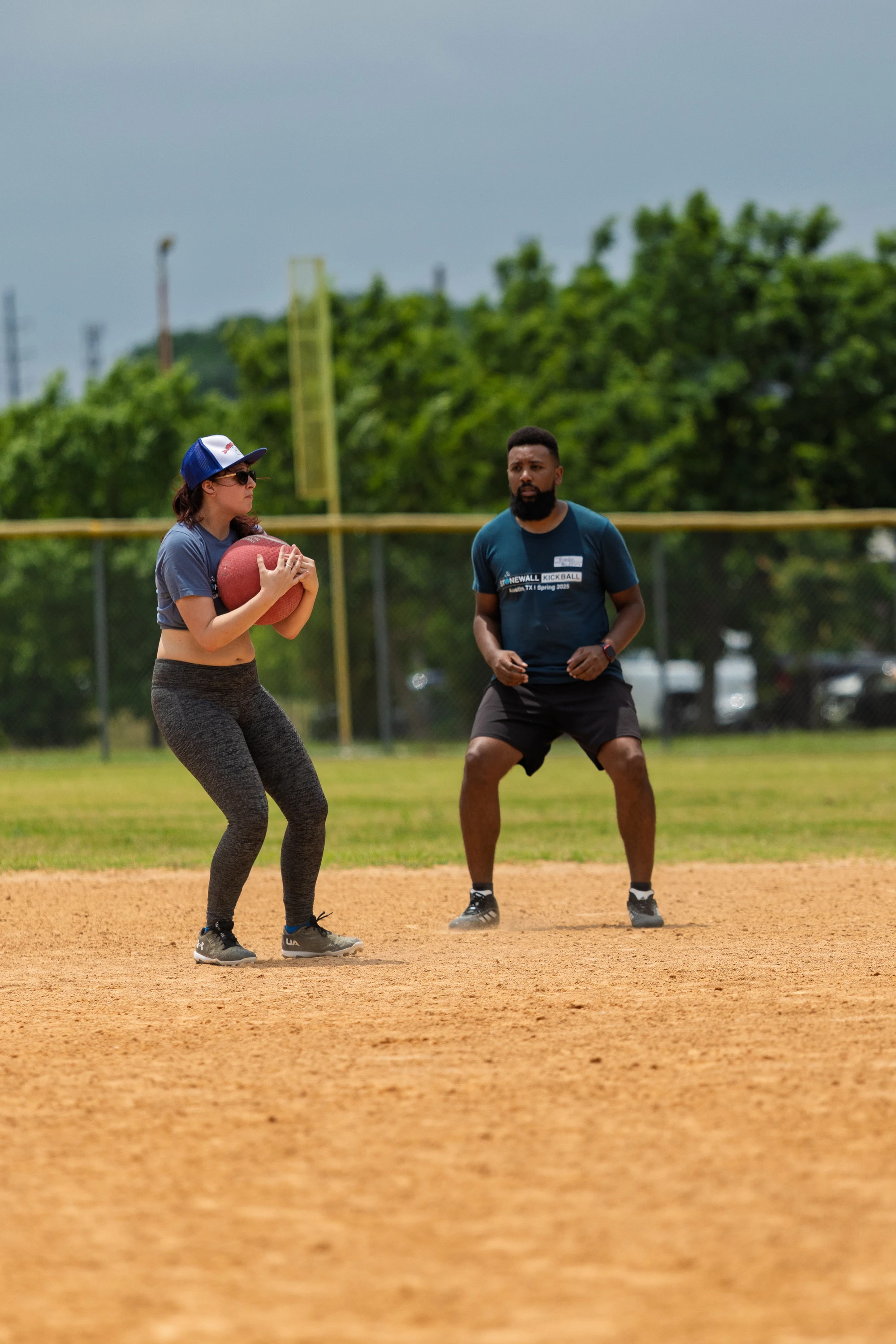 Two kickball players are on a dirt field; one player in a blue cap and athletic clothing is holding a red kickball, while another player in a dark shirt and shorts appears ready to move or defend, with a grassy outfield and trees in the background.