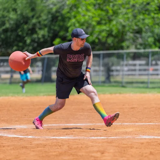 A player wearing a "Pride Kickball" shirt, colorful socks, rainbow wristbands, and a cap is preparing to pitch a kickball on an outdoor field.