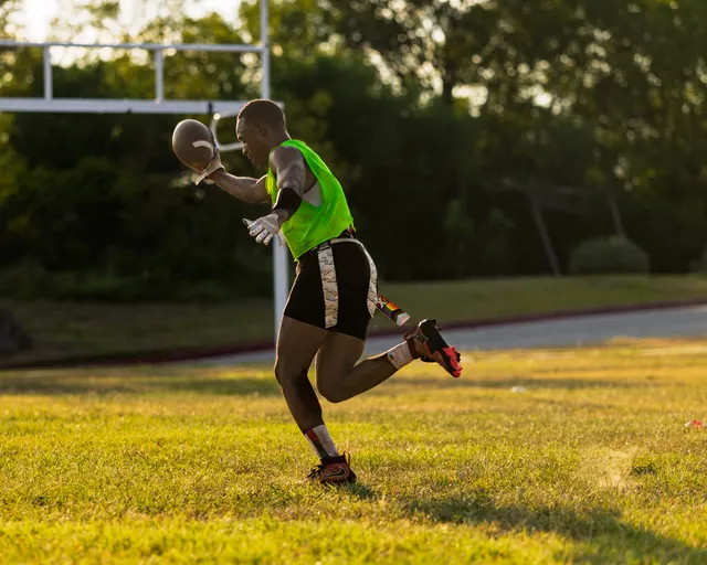 An ATX Heat (Black) player sprints across a sunlit grassy field during flag football practice for Gay Bowl 2025, carrying a football and wearing a neon green top, black shorts with patterned side stripes, gloves, a flag belt, and bright cleats, with a goalpost and trees in the background.