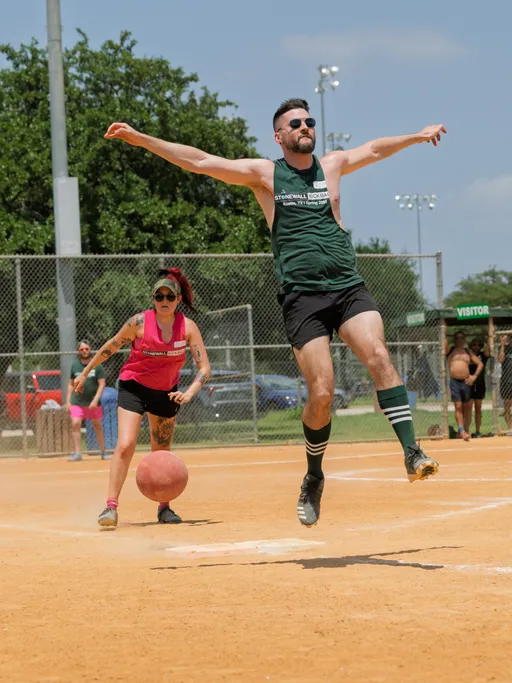 A player wearing a dark green tank top, black shorts, and striped socks leaps in the air after kicking a red kickball, while another player in a pink tank top and black shorts stands nearby on the dirt kickball field. The scene takes place outdoors on a sunny day, with a chain-link fence, trees, and additional players and spectators in the background.