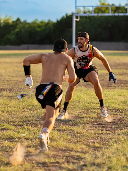 On a dry grass field at sunset, two ATX Heat (Black) flag football players practice for Gay Bowl 2025: a runner with a colorful flag belt and gloves charges forward while a crouched defender in an ATX Heat tank, headband, and cleats braces to pull the flag near the goalposts, dust kicking up between them.