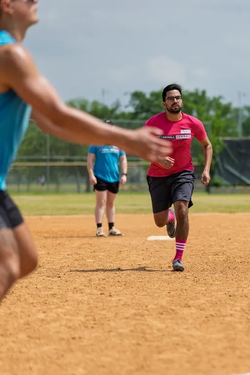 A player wearing a pink "Stonewall Kickball" shirt, black shorts, and striped socks is running on a kickball field, while another player in a blue shirt stands in the background and another competitor runs past in the foreground.