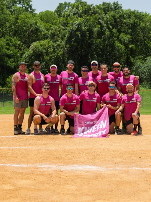 A group of kickball players wearing matching bright pink sleeveless shirts poses together on a dirt kickball field, with some athletes kneeling in front and others standing behind, holding a pink team banner. Lush green trees form the background behind the fence.