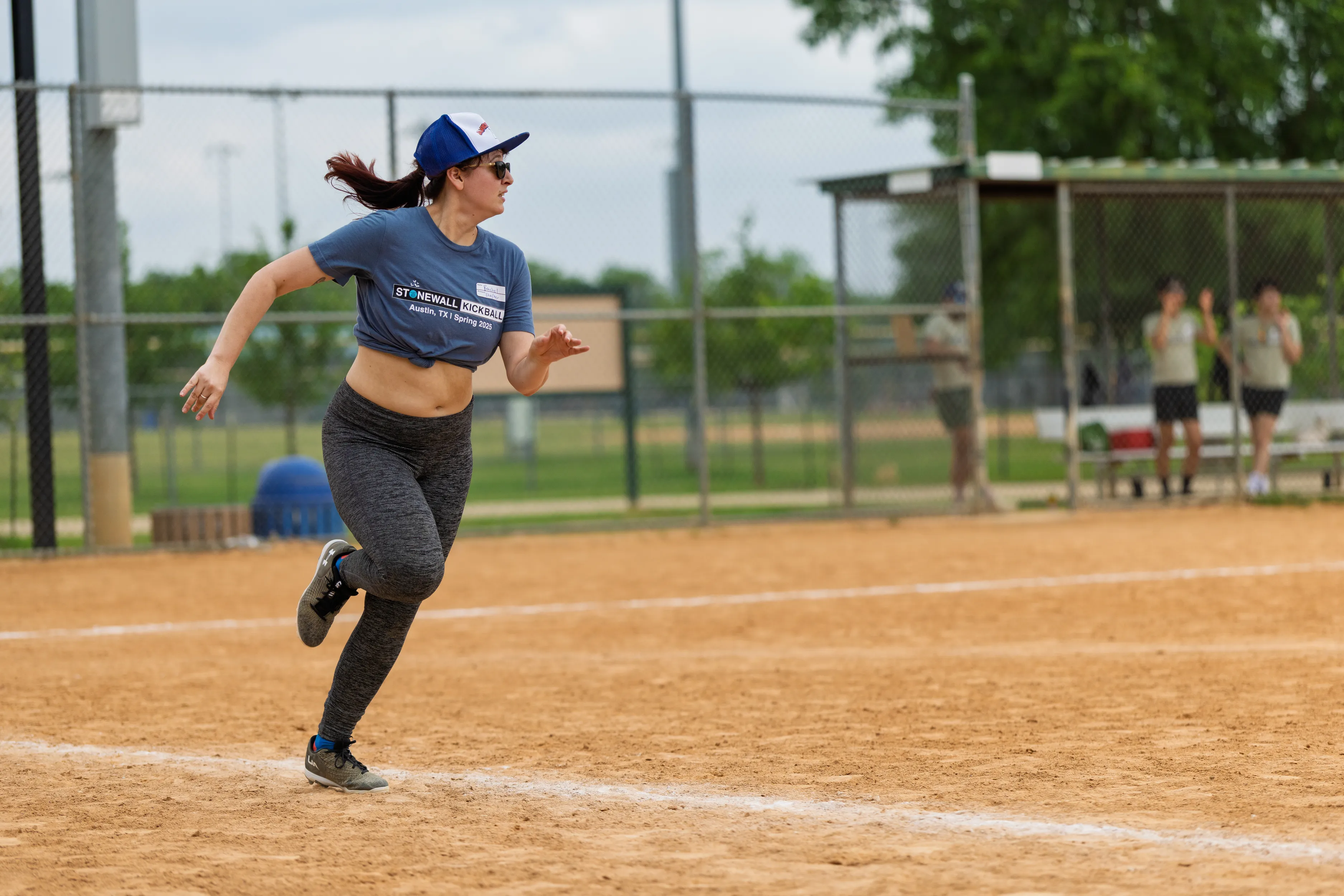 A player sprints along the kickball infield, mid-stride and looking toward the next base, wearing a blue Stonewall Kickball tee, dark leggings, running shoes, sunglasses, and a blue-and-white cap. A chain-link dugout with teammates and benches sits in the background of the park field.