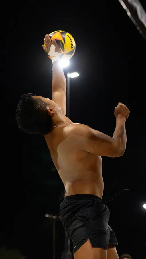 A sand volleyball player in black shorts leaps at night under bright lights, extending one arm with a taped wrist to strike a multicolored ball near the net. The action unfolds on an outdoor sand court, with sand kicking up as the athlete jumps.