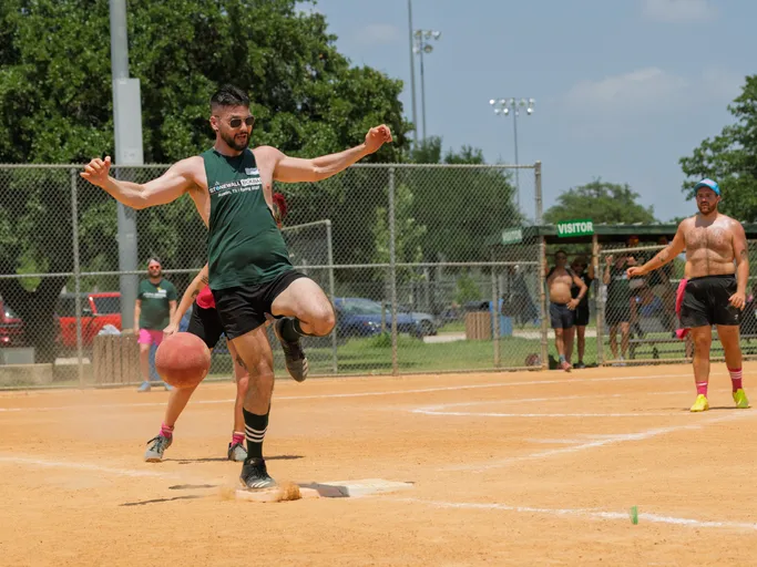 A player in a dark sleeveless shirt and shorts is kicking a red kickball on a sandy field, while other players and spectators watch from behind a chain-link fence and near a dugout labeled "Visitor."