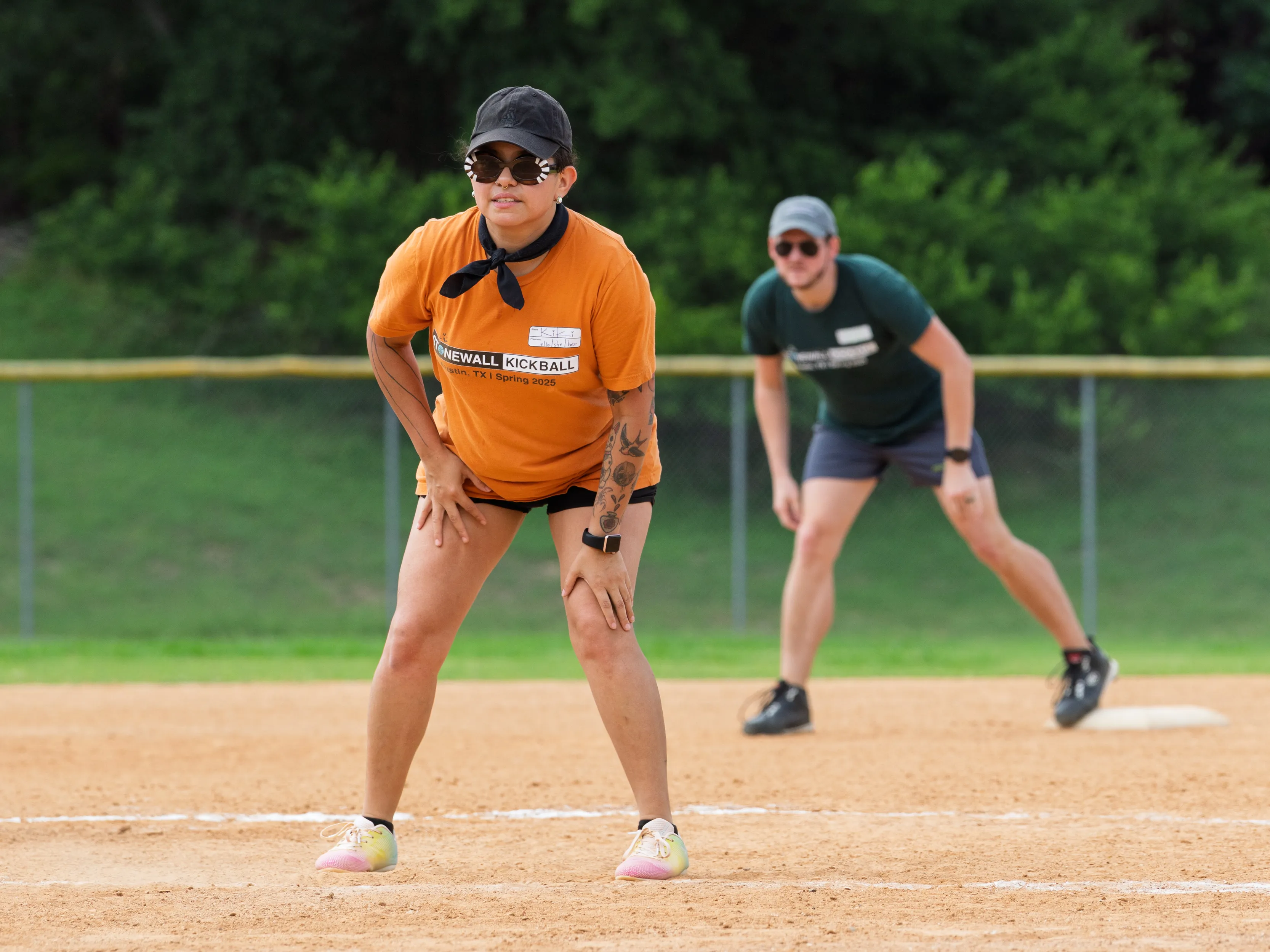 A player in an orange Stonewall Kickball shirt, black shorts, cap, sunglasses, and a neck bandana crouches on the infield with hands on knees, ready for the next play. Another player in a green shirt prepares behind near a base on a dirt kickball field bordered by grass, a chain-link fence, and trees.
