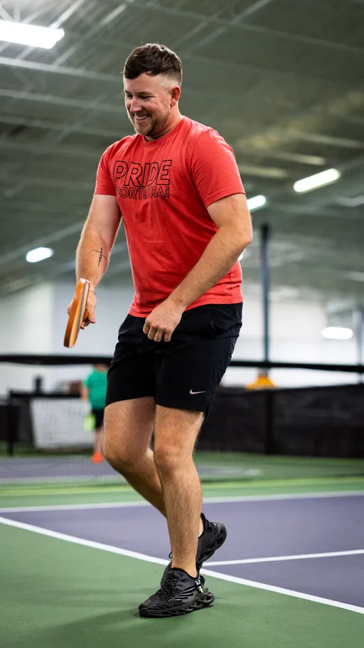 A pickleball player in a red "Pride Sports USA" shirt and black shorts is actively moving on an indoor pickleball court, holding a paddle and appearing focused on the game. The setting is a well-lit sports facility with other players and courts visible in the background.