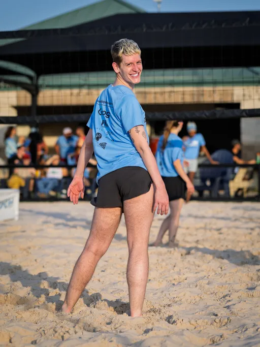 A volleyball player stands on a sandy court, smiling toward the camera, wearing a light blue team shirt and black shorts, with other players and spectators visible in the background.