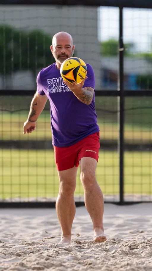 A volleyball player wearing a purple "PRIDE SPORTS USA" shirt and red shorts prepares to serve a yellow and black volleyball on a sand court, with a metal fence and grassy area visible in the background.