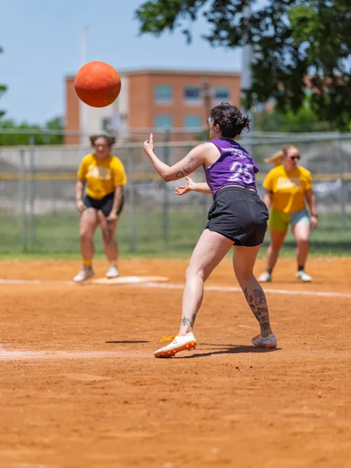 A kickball player in a purple jersey and black shorts is preparing to catch a kickball while running on a dirt field; several competitors in yellow shirts are visible in the background, and a building can be seen beyond the field.