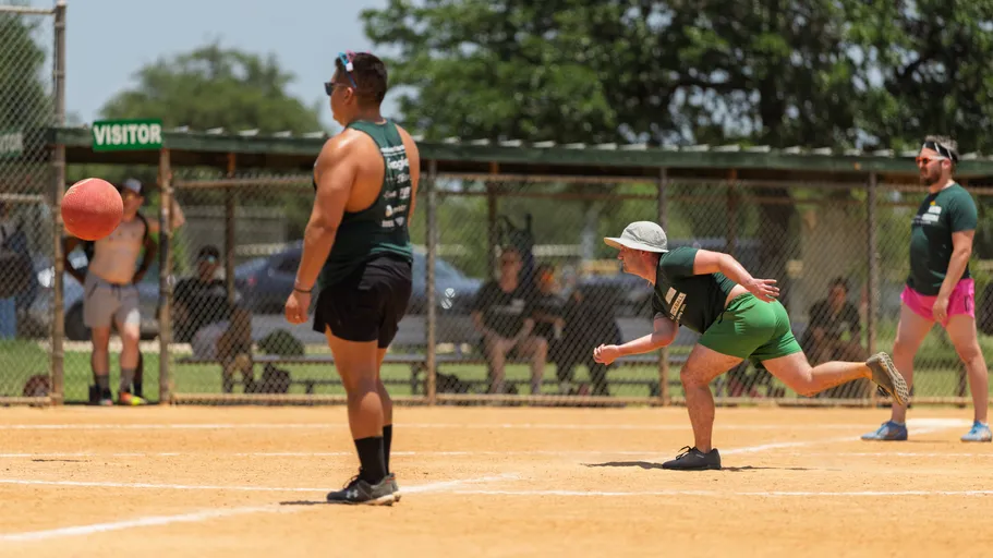 A player wearing a sun hat and green athletic outfit is rolling a red kickball towards home plate, while other players in athletic clothing stand ready on the dusty kickball field with a chain-link fence and spectators in the background.
