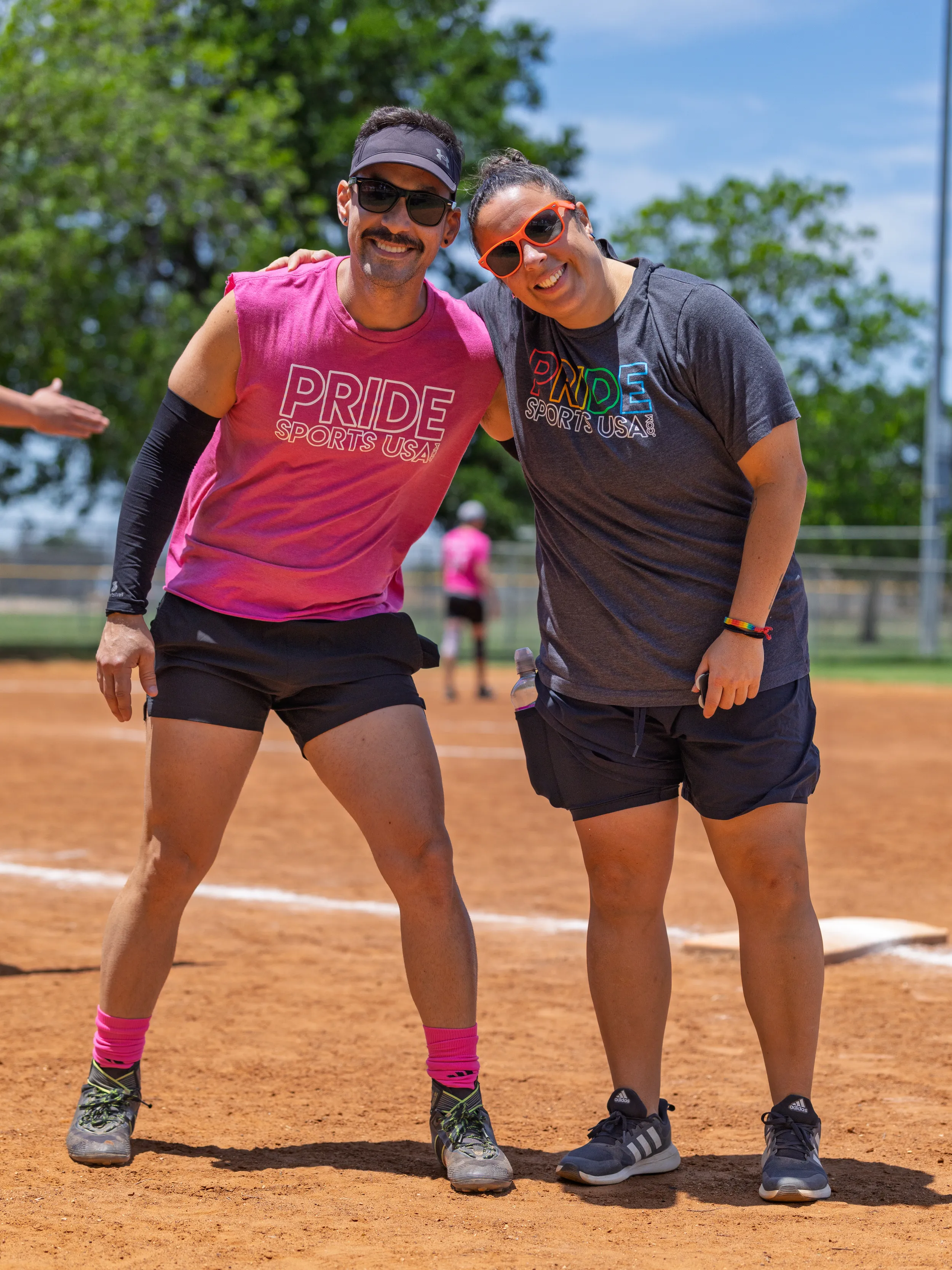 Two players pose together on a sunny kickball field, wearing “PRIDE SPORTS USA” shirts—one in a pink sleeveless top with a visor, sunglasses, arm sleeve, black shorts, and bright pink socks; the other in a dark gray tee with rainbow lettering, orange sunglasses, navy shorts, and a wristband. They stand on the dusty infield near the baseline, with trees, a fence, and other players blurred in the background.