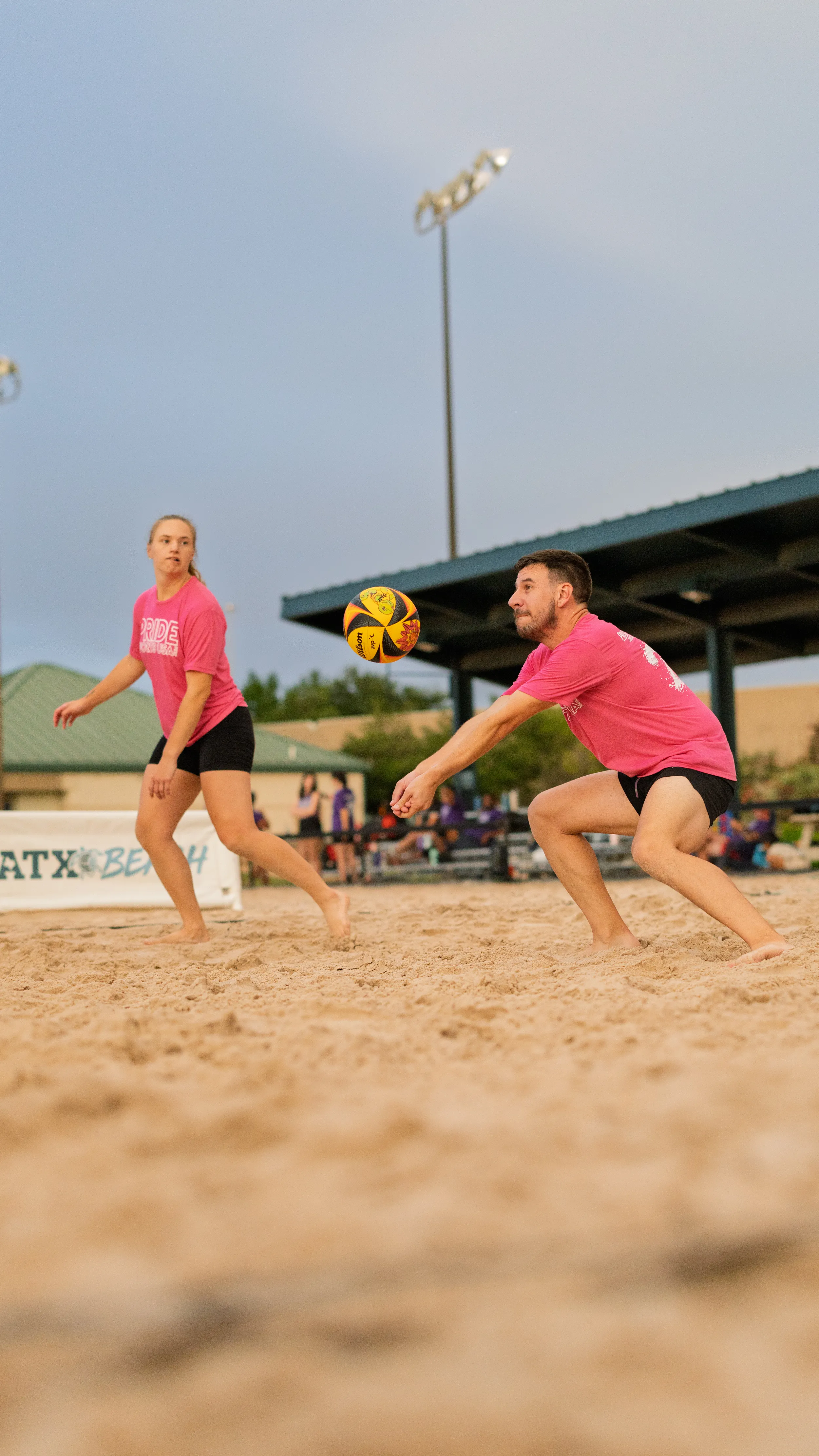 Two sand volleyball players in matching pink Pride shirts and black shorts compete on an outdoor court at dusk; one drops into a low forearm pass to bump a yellow-and-black ball while a teammate tracks the play, with lights, pavilion, and ATX Beach signage in the background.