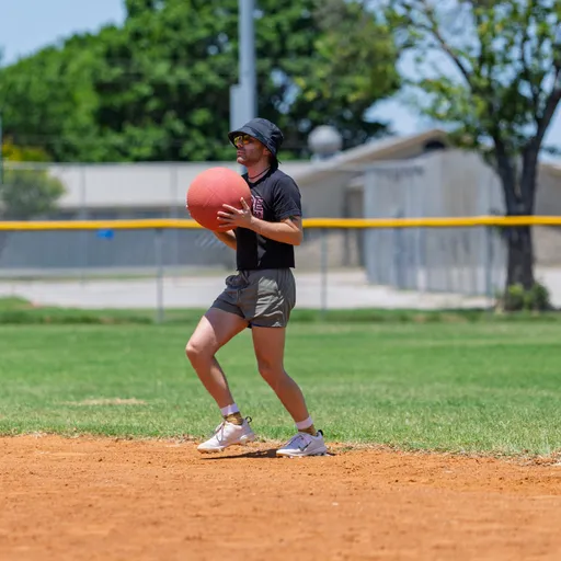 A kickball player in athletic shorts, a bucket hat, and a dark t-shirt prepares to throw a red kickball on a sunny outdoor field surrounded by grass and a yellow fence.