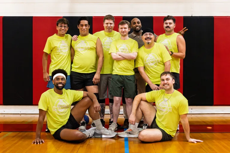 Nine dodgeball players in matching neon yellow team shirts, athletic shorts, and sneakers pose together in an indoor gym with a wood floor and red-and-black wall padding; two kneel in front while the others stand close behind, some with arms around teammates, and a few wear a headband or cap.
