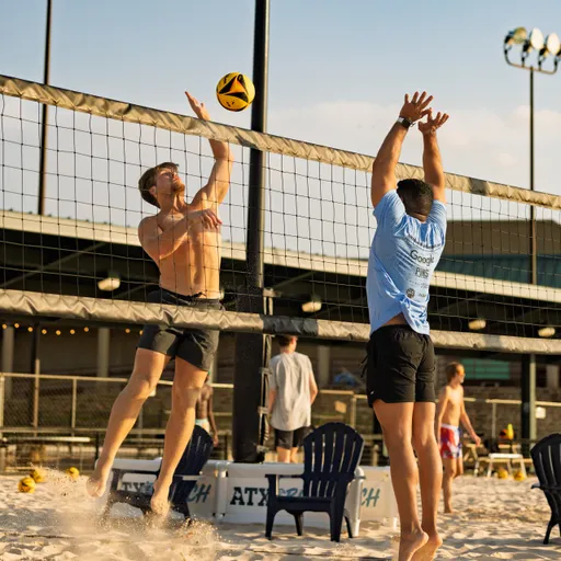 Two sand volleyball players jump at the net, reaching up to block or spike the ball, wearing athletic shorts and shirts, with additional players and spectators visible in the sandy outdoor court setting.