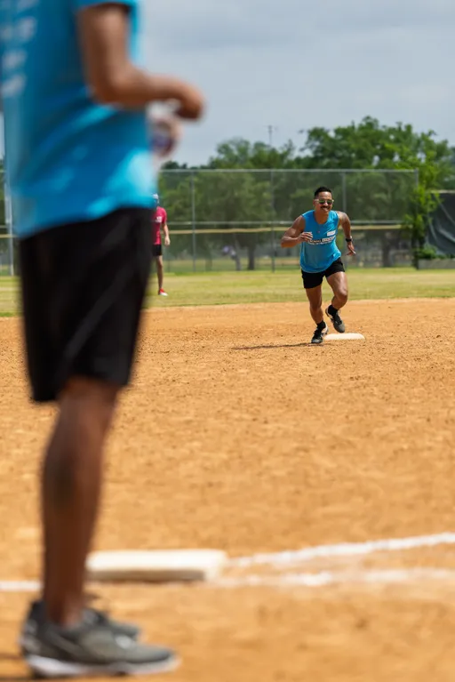 A kickball player in a blue tank top and black shorts is running from a base on a dirt kickball field, while another player stands in the foreground wearing similar athletic clothing and sneakers. A chain-link fence and grassy area are visible in the background.