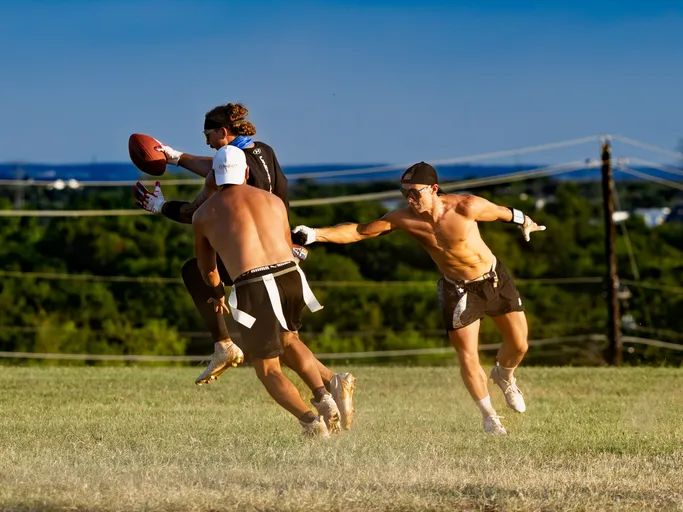 At an ATX Heat (Black) flag-football practice for Gay Bowl 2025, a receiver in a black long-sleeve top, white gloves, blue headband, and gold cleats leaps to secure the ball while two defenders in shorts and flag belts converge, one reaching for a flag. The play unfolds on a sunlit grassy field with dust kicking up, tree-covered hills, and power lines in the background.