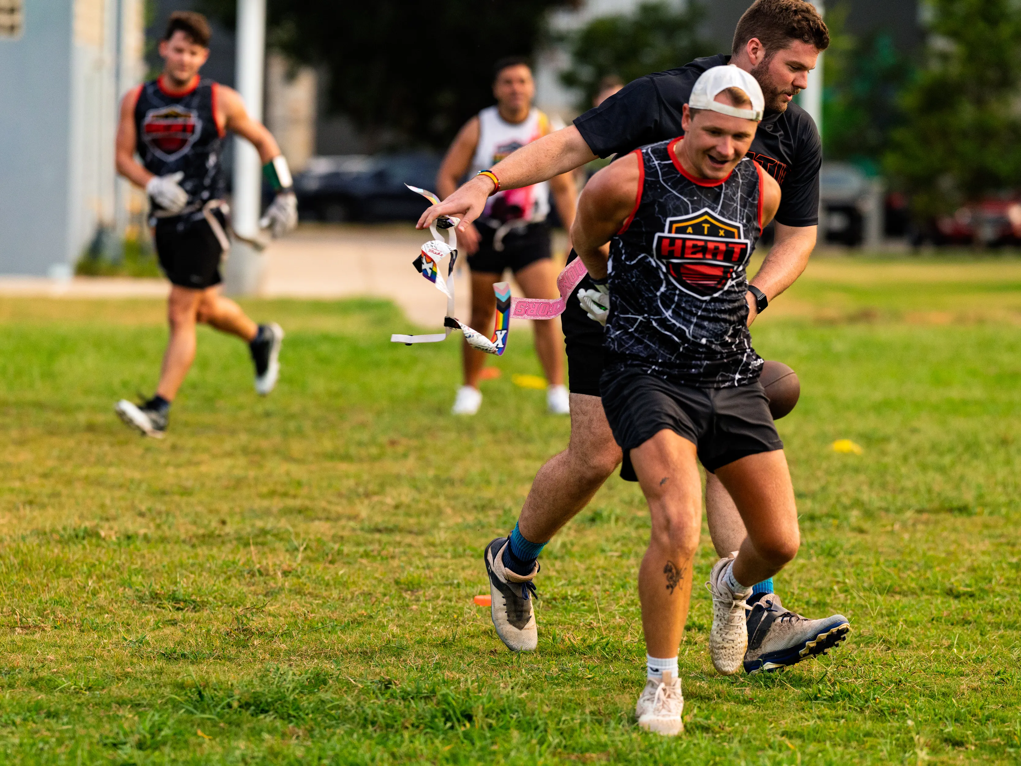 On a grassy flag football field, ATX Heat (Orange) players run drills as a defender yanks a runner’s flag mid-sprint; athletes wear black ATX Heat jerseys with orange trim, flag belts, shorts, cleats, and a backward cap, with cones marking the practice area.