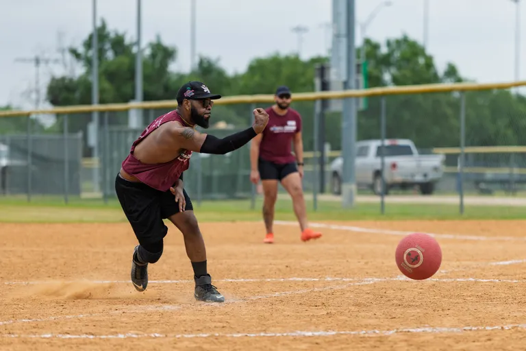 A kickball player wearing a maroon jersey, black shorts, a black cap, and an arm sleeve is mid-pitch on a dirt field, while another player in similar attire stands in the background near the pitcher's mound.
