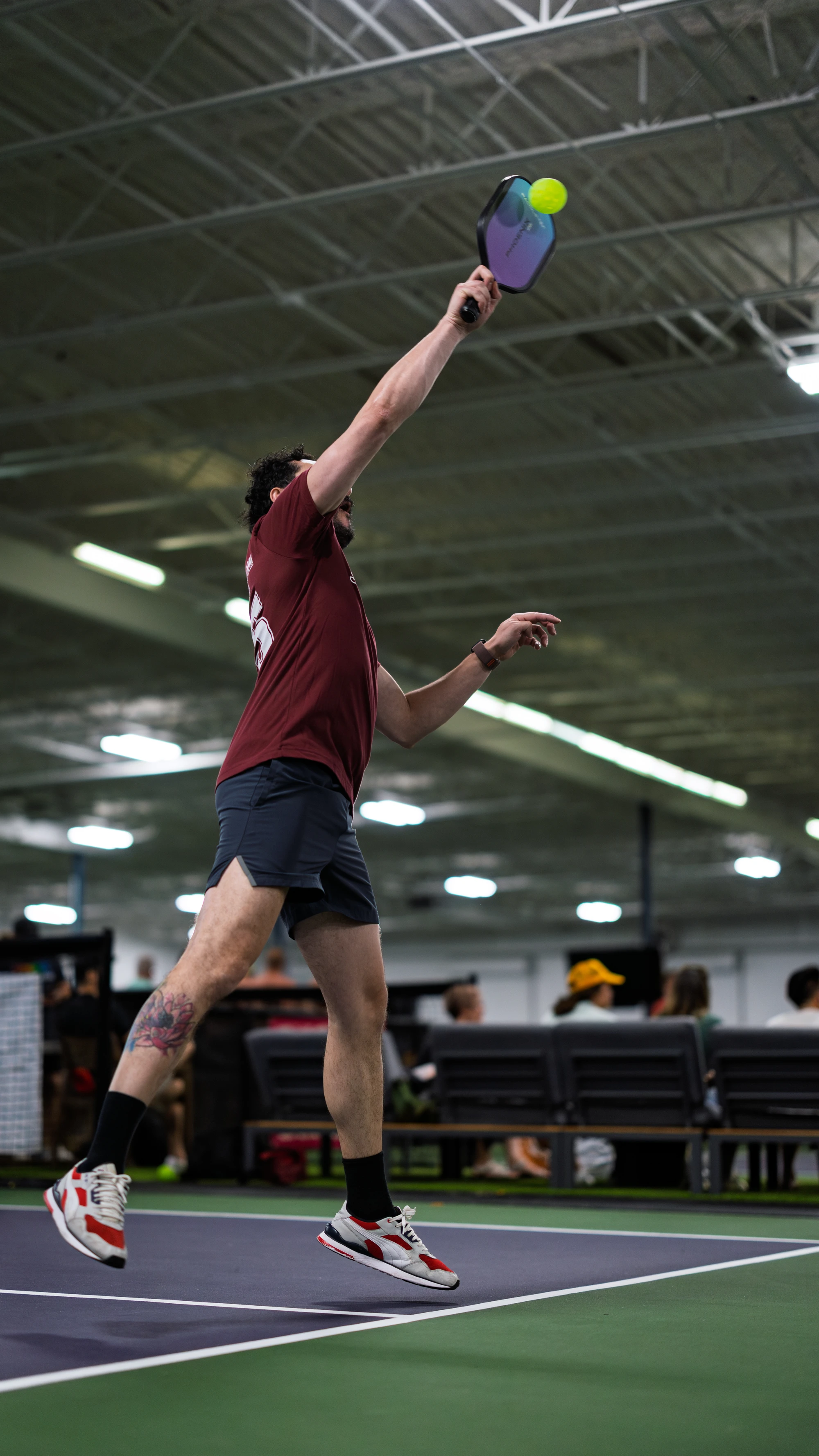 A pickleball player in athletic clothing jumps to hit a ball with a paddle on an indoor court, with spectators seated in the background.
